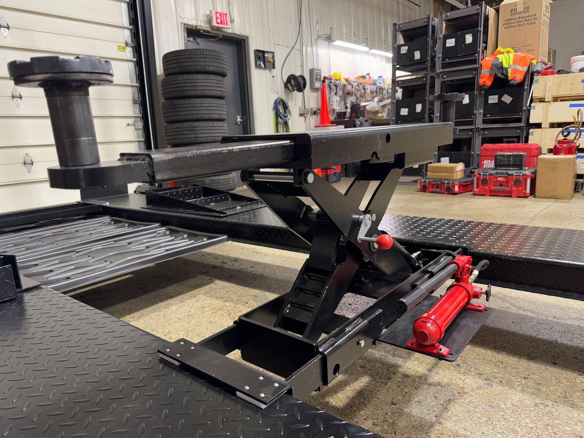 Black hydraulic vehicle lift in an auto shop with a red floor jack and tire stacks in the background