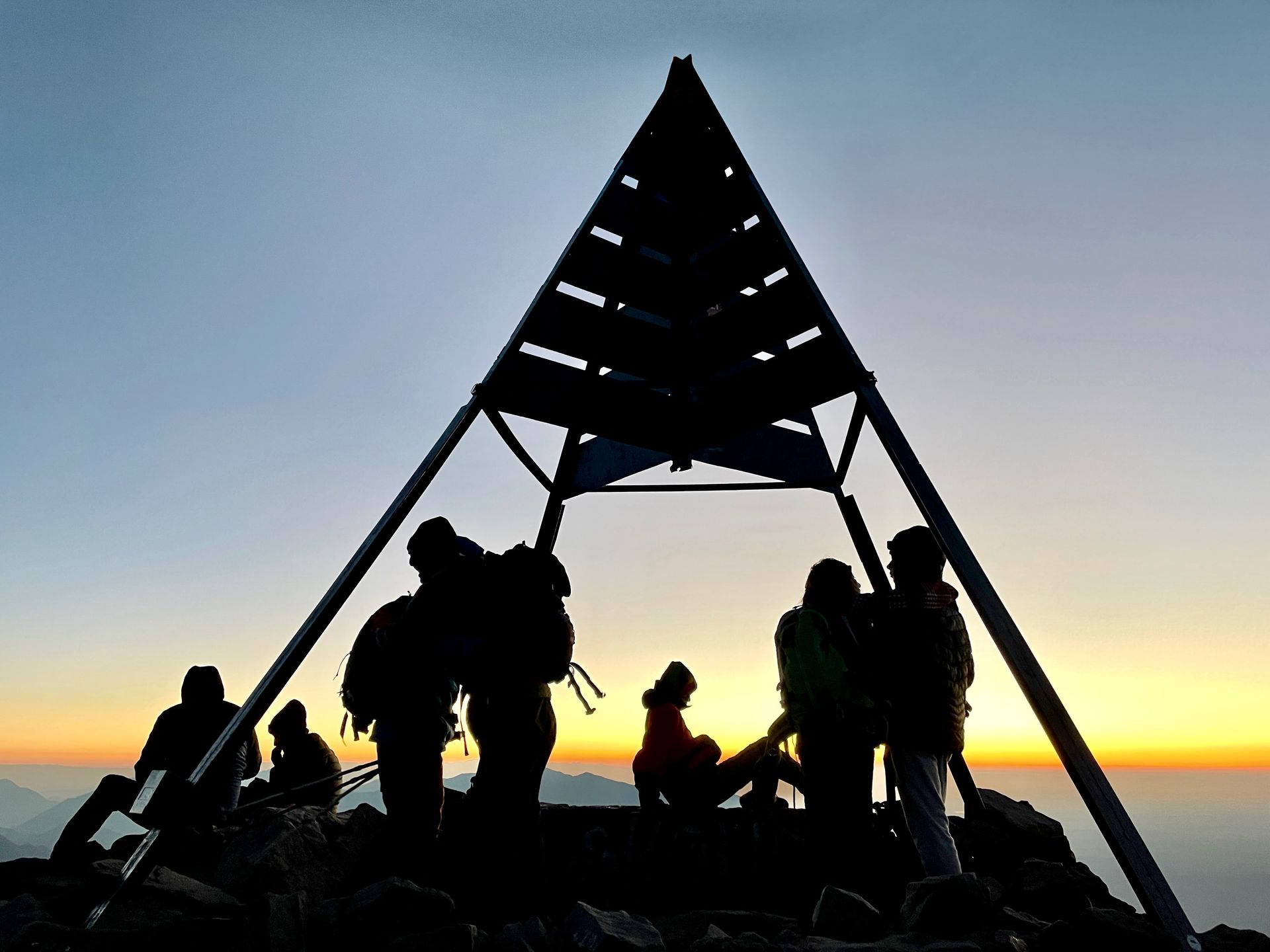 Silhouetted hikers gather under a triangular lookout at sunset with a blue-orange sky.