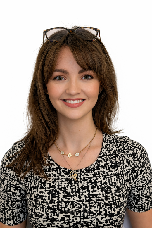 A smiling person with brown hair and bangs, wearing glasses atop their head, a patterned shirt, and a layered necklace.