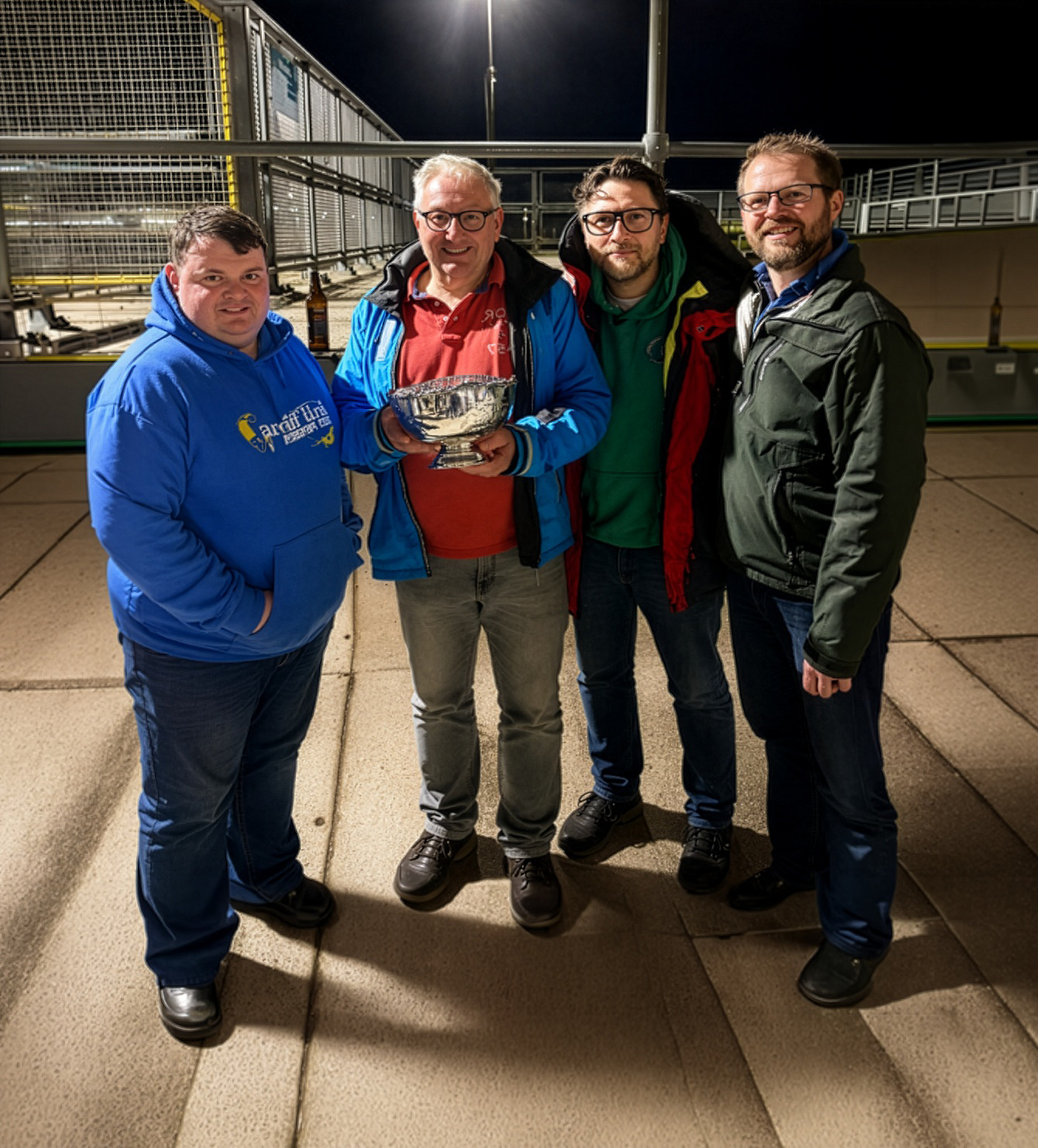 Four people standing by a fence at night, one holding a fish, all smiling at the camera.