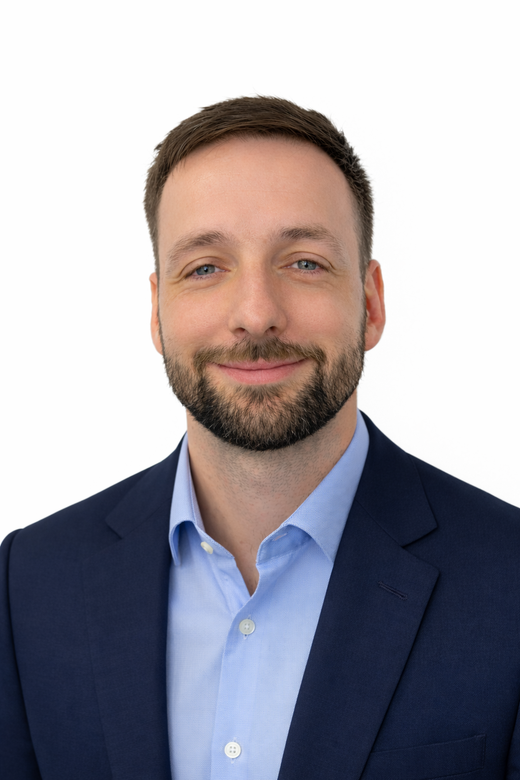 Headshot of a man with short brown hair and a beard, wearing a light blue button-down shirt and a navy blazer.