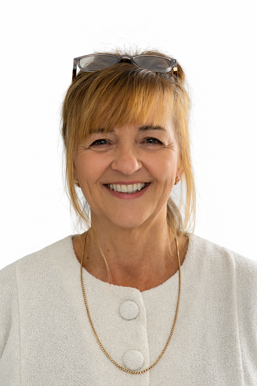 A smiling woman with blonde hair, wearing a white textured top, glasses on her head, and a thin gold chain necklace.
