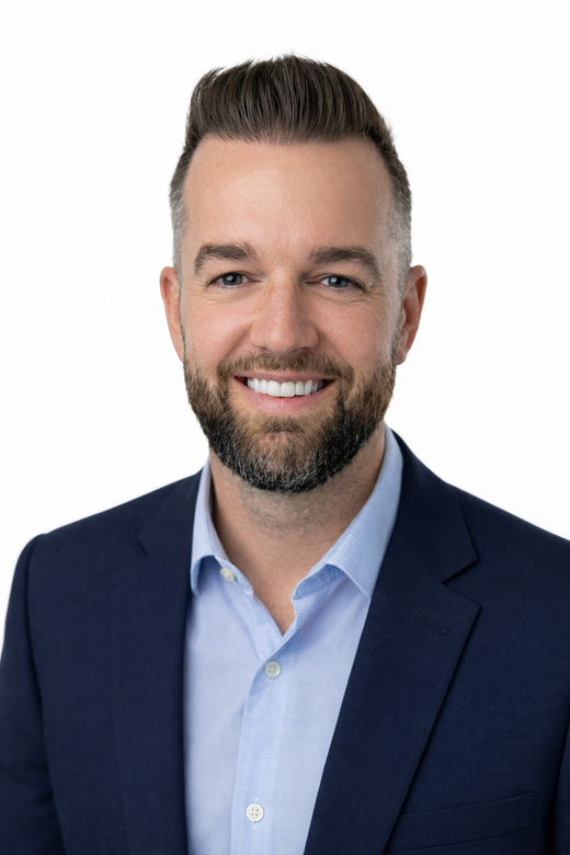 A professional headshot of a smiling person with a groomed beard, wearing a light blue dress shirt and a navy blue blazer.