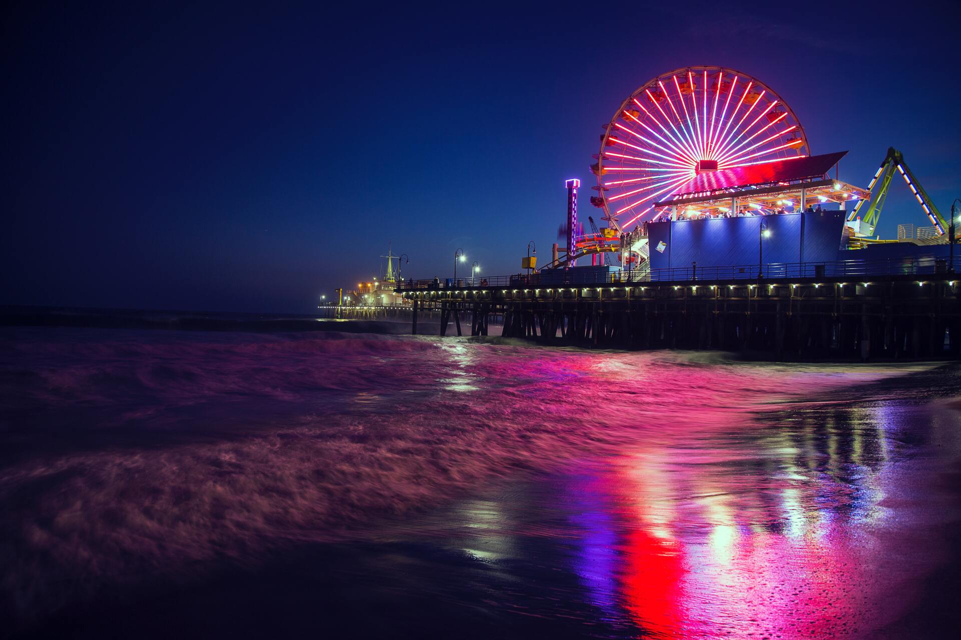 A ferris wheel is lit up at night on a pier overlooking the ocean.