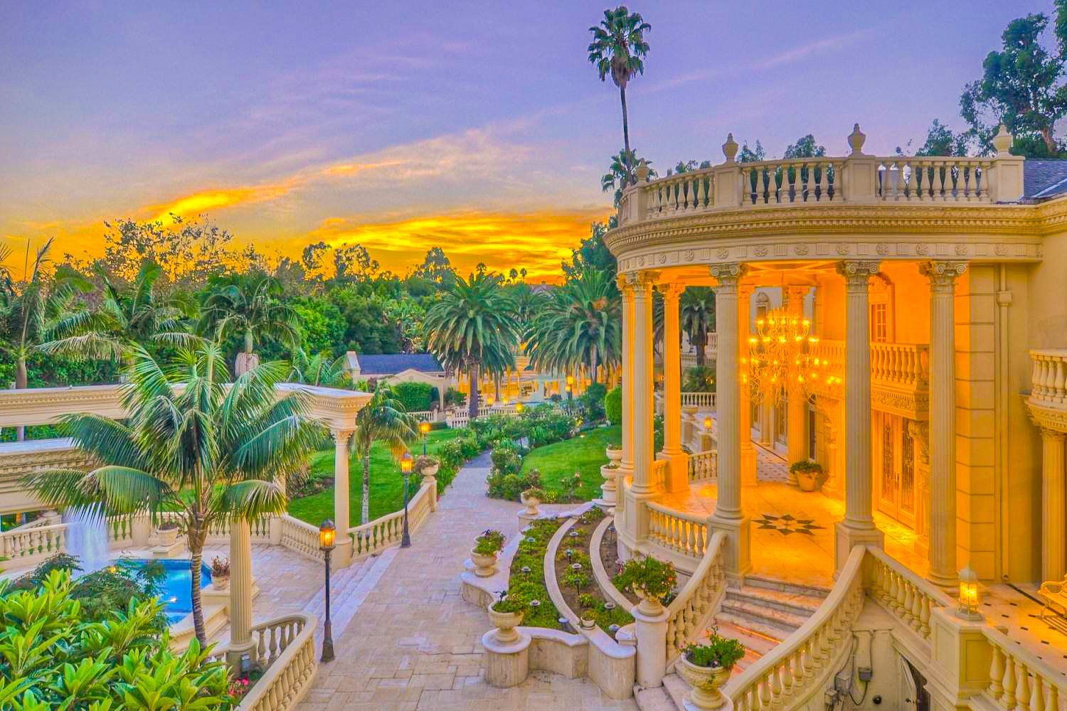 An aerial view of a large mansion with a balcony at sunset surrounded by palm trees.