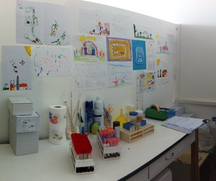 Banco da laboratorio con provette e materiali di consumo sotto colorati disegni infantili sulla parete.