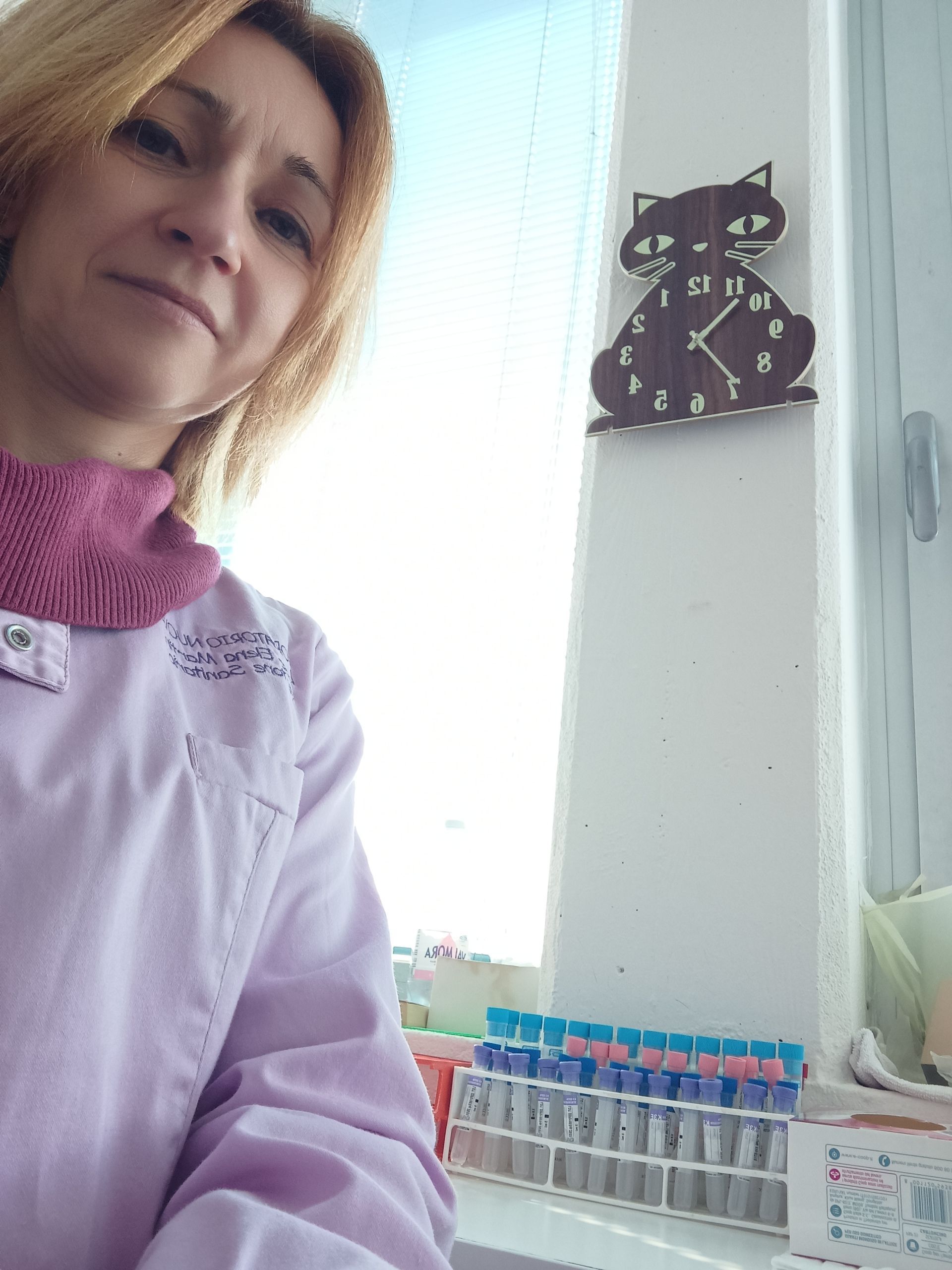 Una donna con un maglione color lavanda è seduta accanto a una finestra luminosa, con una decorazione a forma di gatto nero sulla parete.