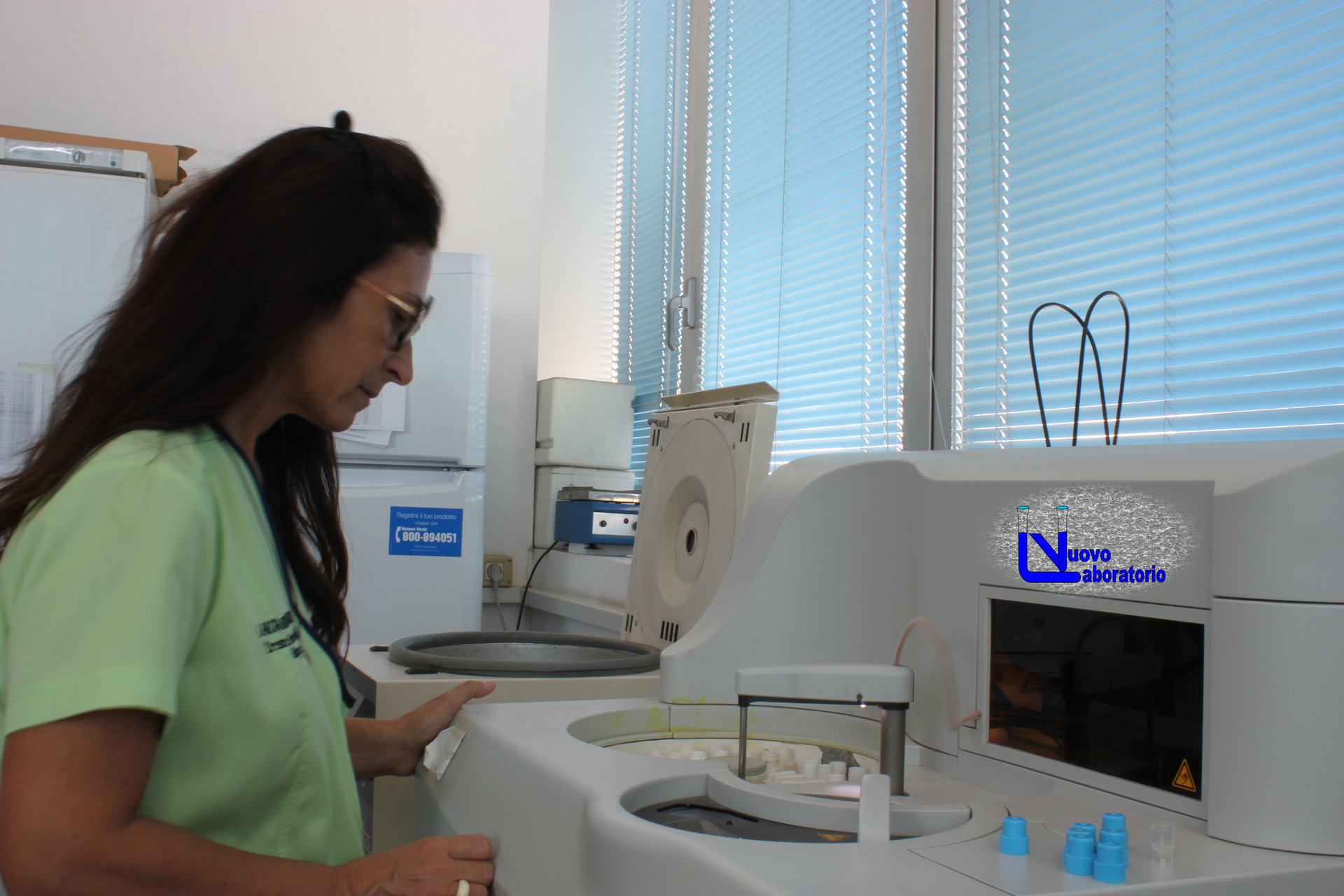 Tecnico con camicia verde che ispeziona le apparecchiature di laboratorio vicino a una finestra con persiane