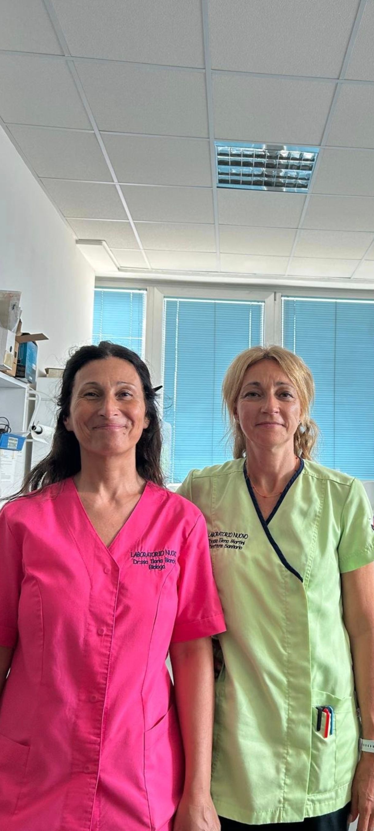 Due persone in camici rosa e verdi in piedi in un luminoso corridoio di una clinica.