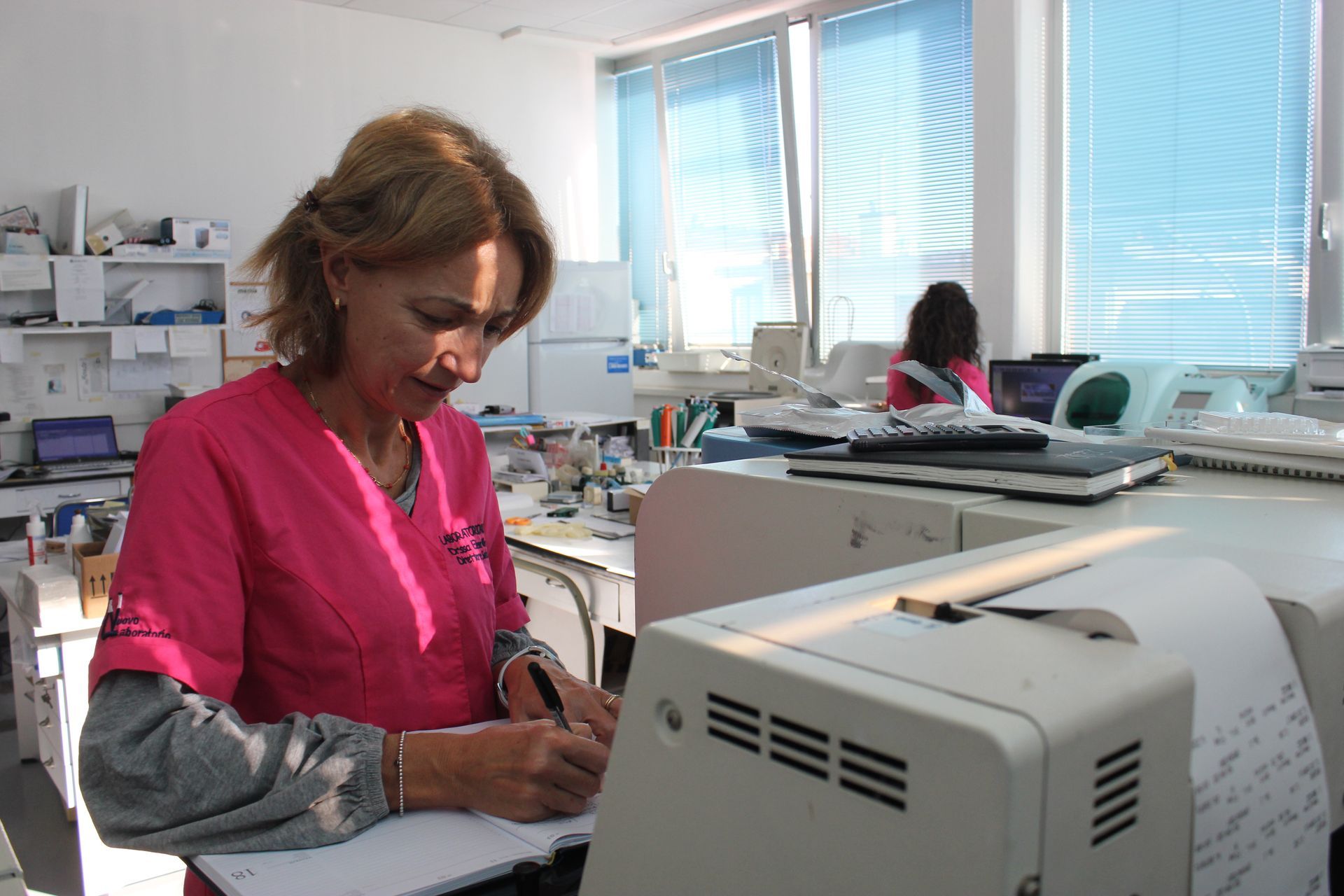 Una donna in camice rosa scrive a una scrivania in un luminoso studio medico.