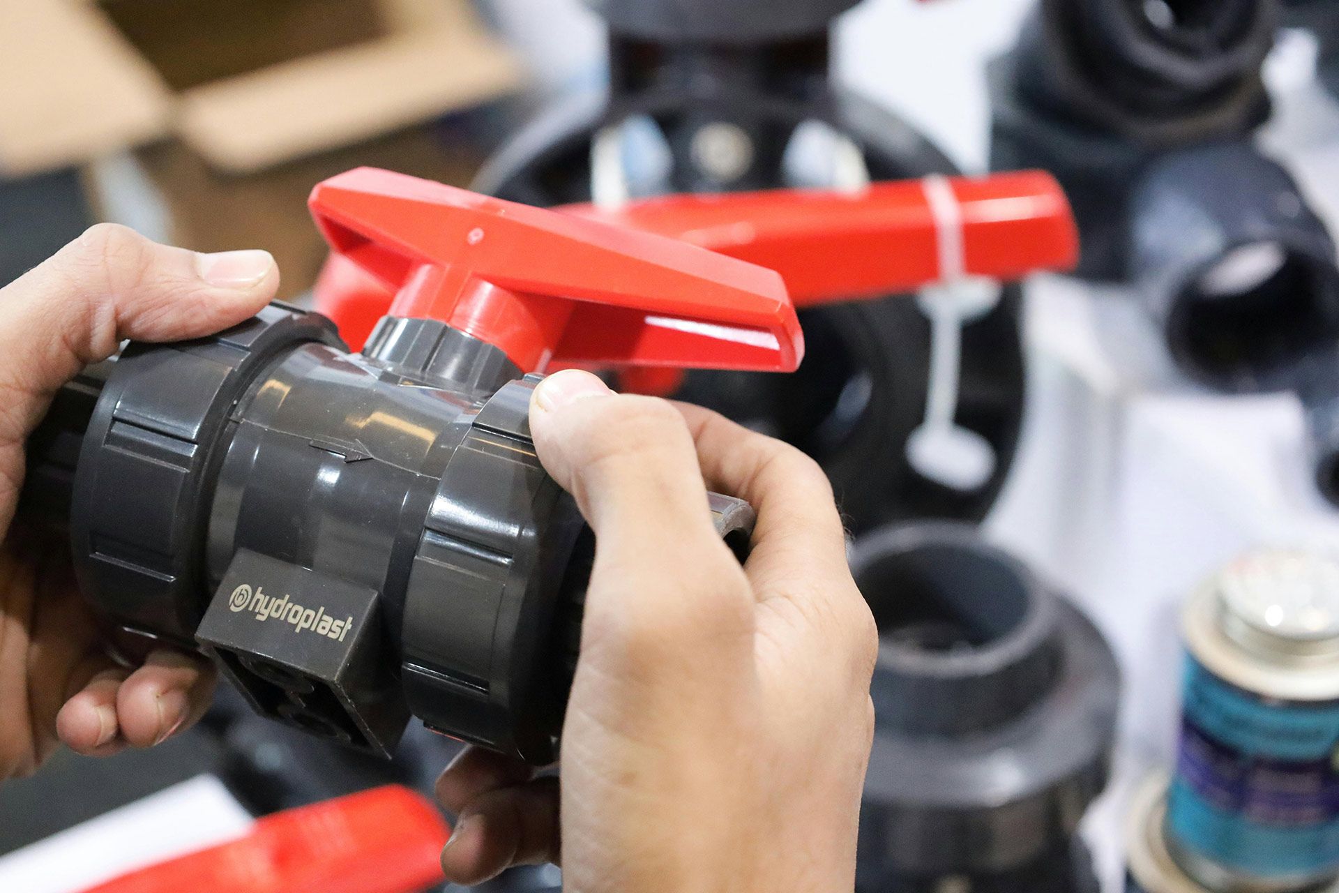 Hands holding a dark gray PVC ball valve with a red handle. Other plumbing parts are in the background.