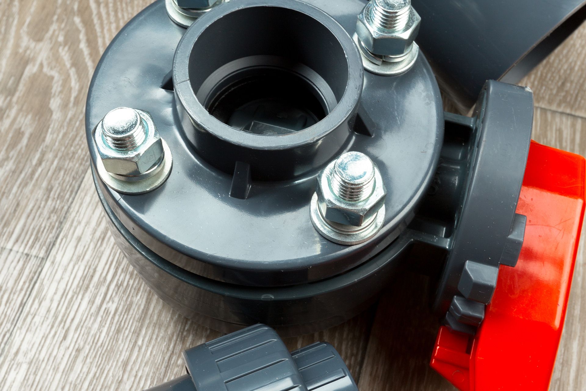 Close-up of a dark gray PVC ball valve with a red handle and multiple bolts; rests on wood.