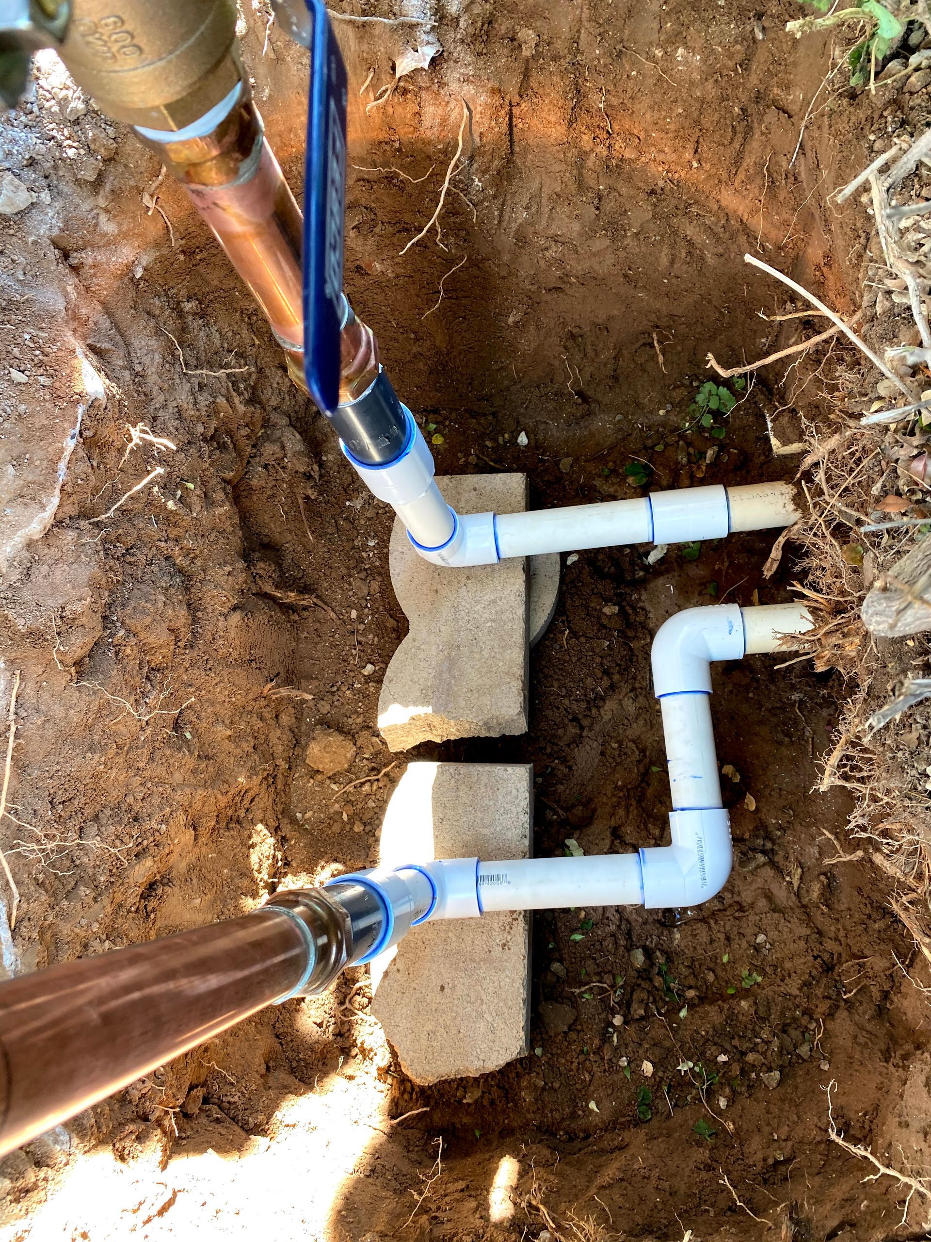 Two white PVC pipes with copper connections and valves are installed in a dirt trench, supported by concrete blocks.