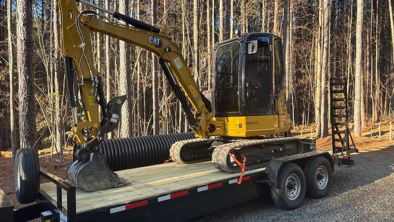 Yellow excavator on a flatbed trailer in a wooded area, with another excavator arm visible nearby.