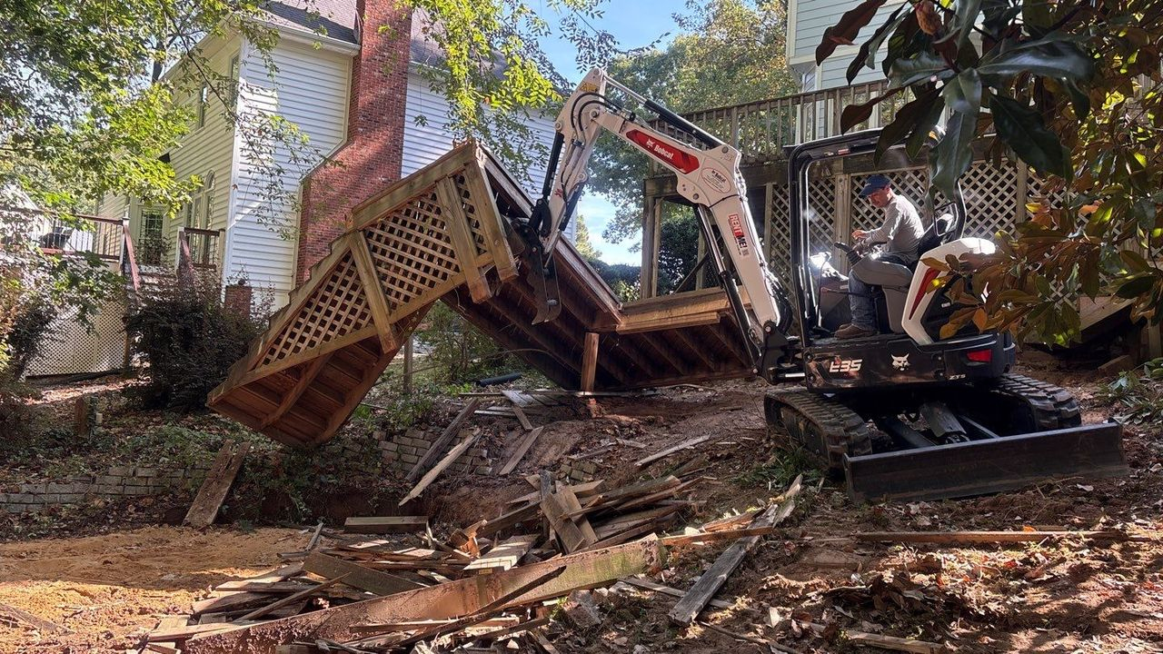 Mini excavator topples a wooden deck in a wooded yard, with debris scattered around.