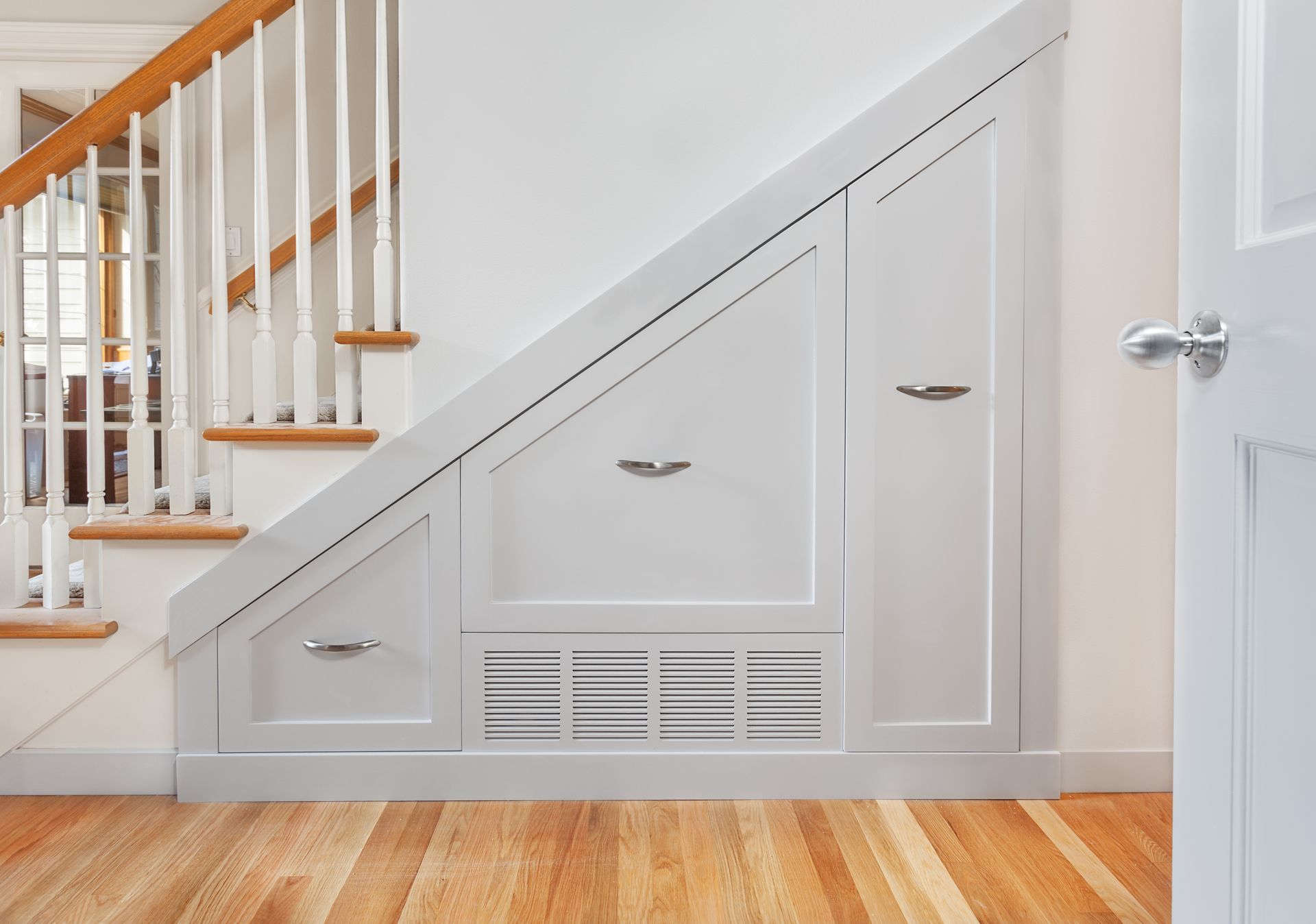 Under-stair built-in storage cabinets in a bright hallway with hardwood floors and white staircase railings