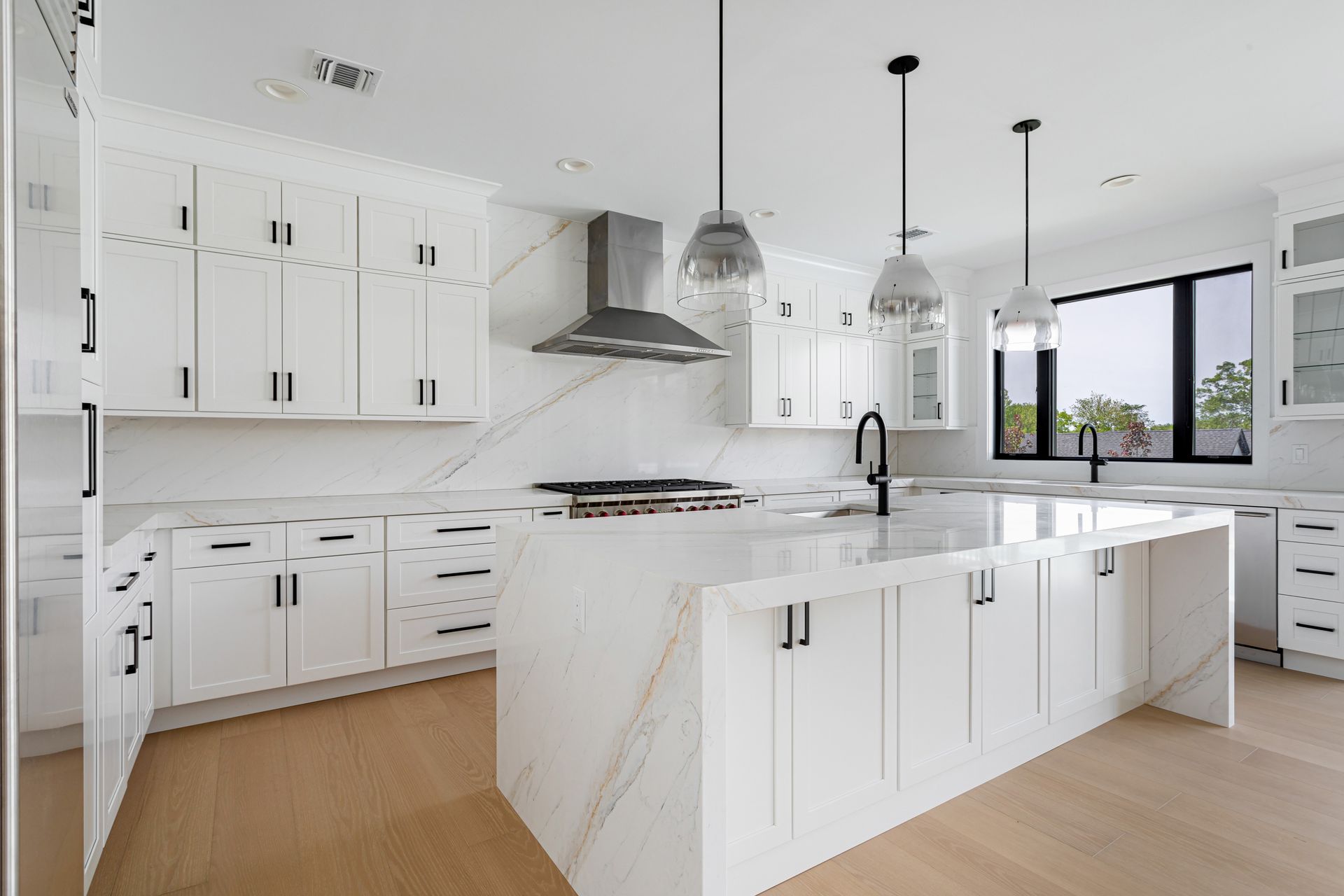 Bright modern white kitchen with marble island, pendant lights, stainless hood, and large windows