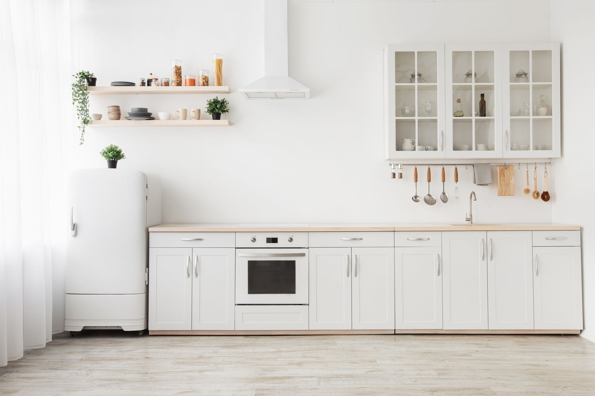 Bright minimalist white kitchen with cabinets, oven, shelves, and hanging utensils