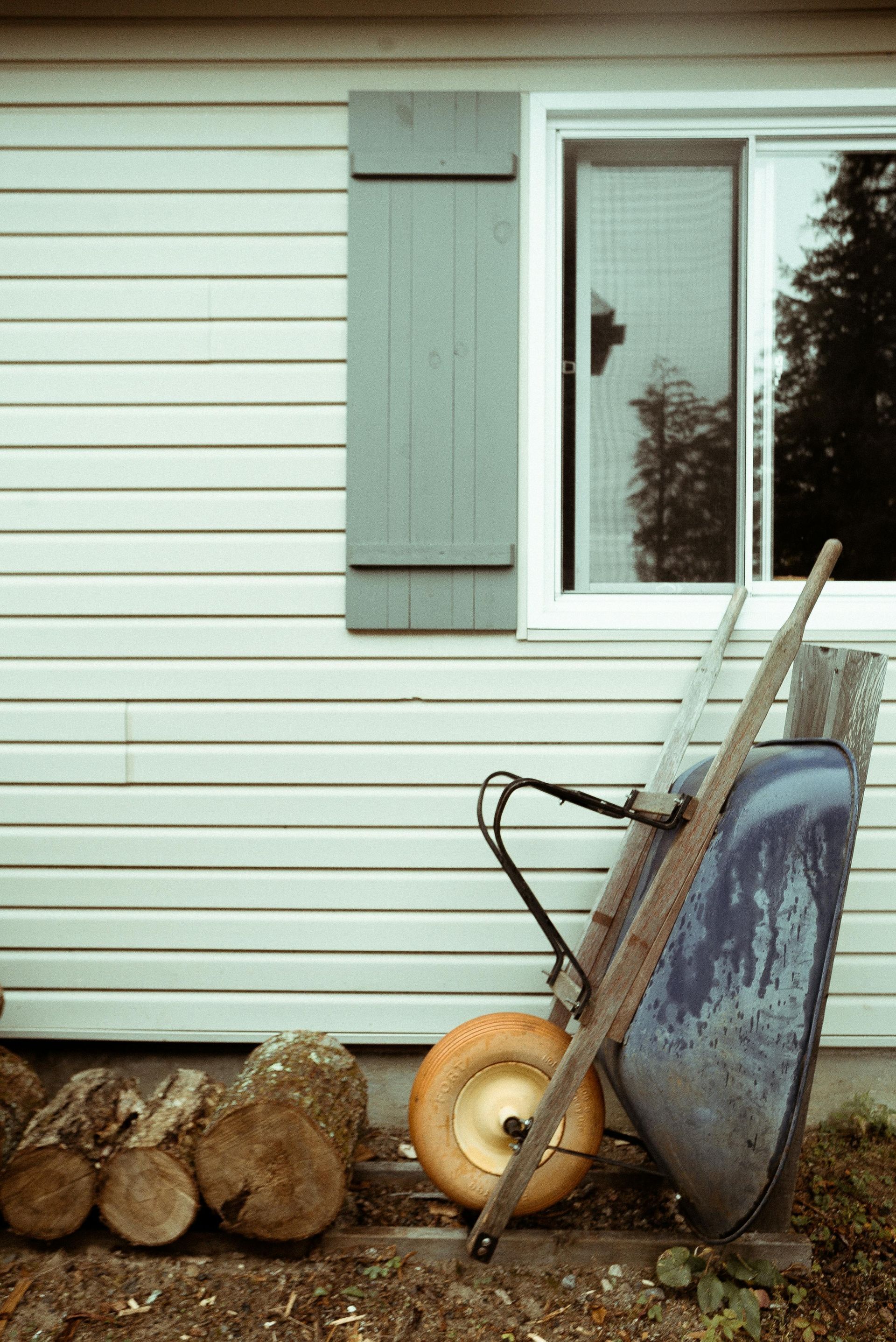 Wheelbarrow next to house with logs and shutter. The wheelbarrow has a yellow tire and is leaning against the house.