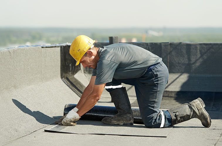 Roofer in hard hat kneels on flat roof, applying dark material. Sunny outdoor setting.