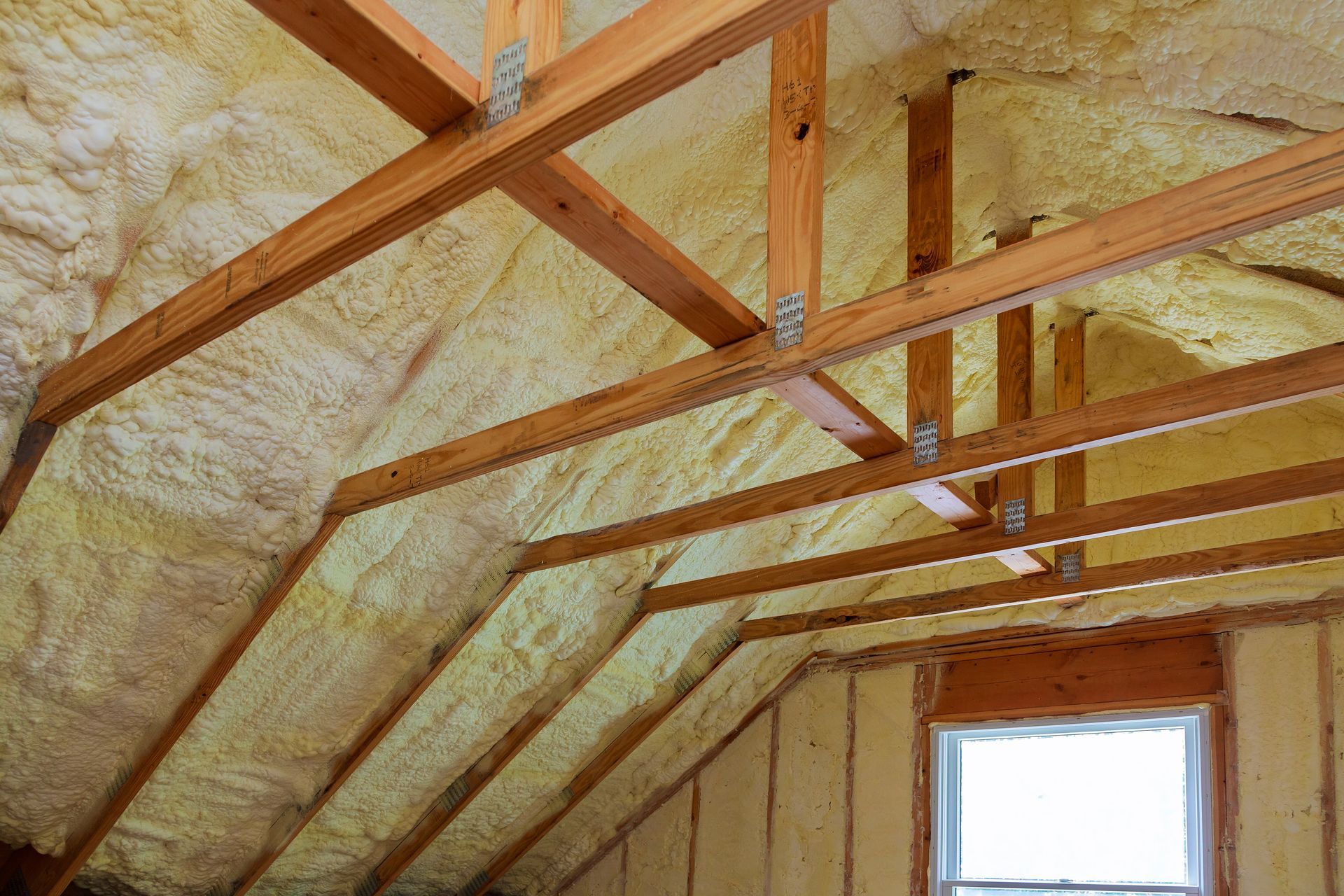 Attic insulation with exposed wooden beams and a small window. Beige spray foam covers the interior.