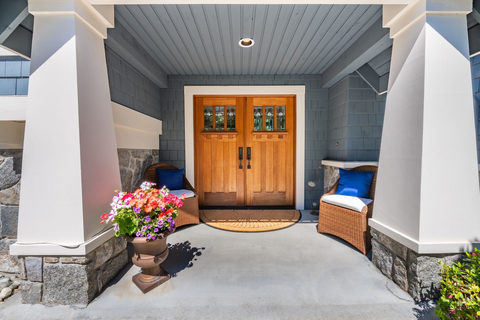 Wooden double doors of a house, flanked by white pillars, stone accents, wicker chairs, and a flower pot.