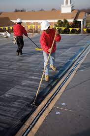 Two roofers in red shirts on a flat roof, applying sealant with long-handled tools.