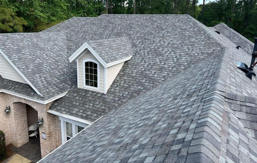 Grey asphalt shingle roof on a house, with a small dormer and trees in the background.