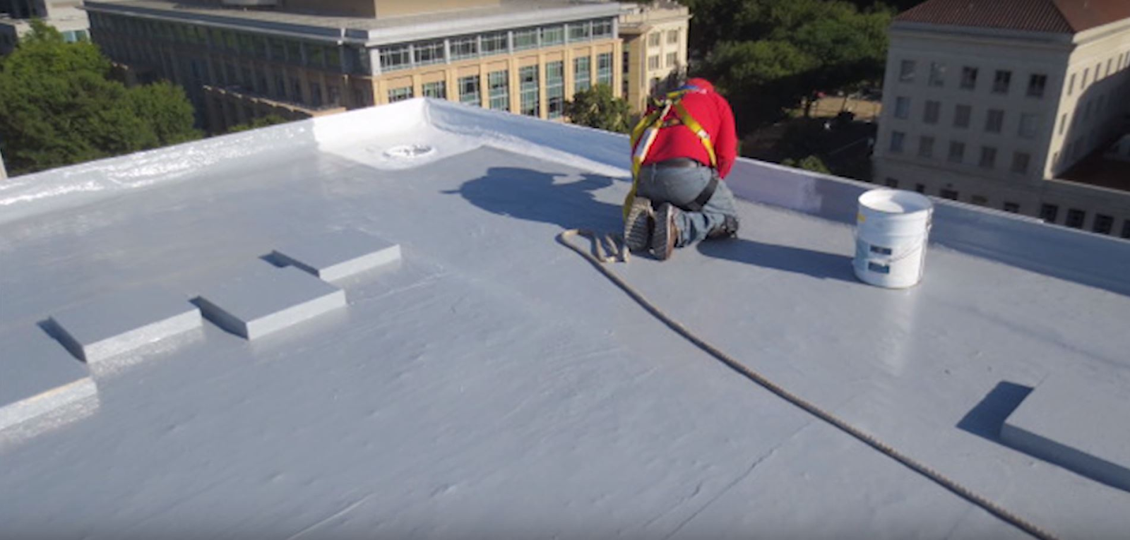 Worker in red kneels on flat gray roof, applying coating with brush. A bucket's nearby and city buildings in the background.