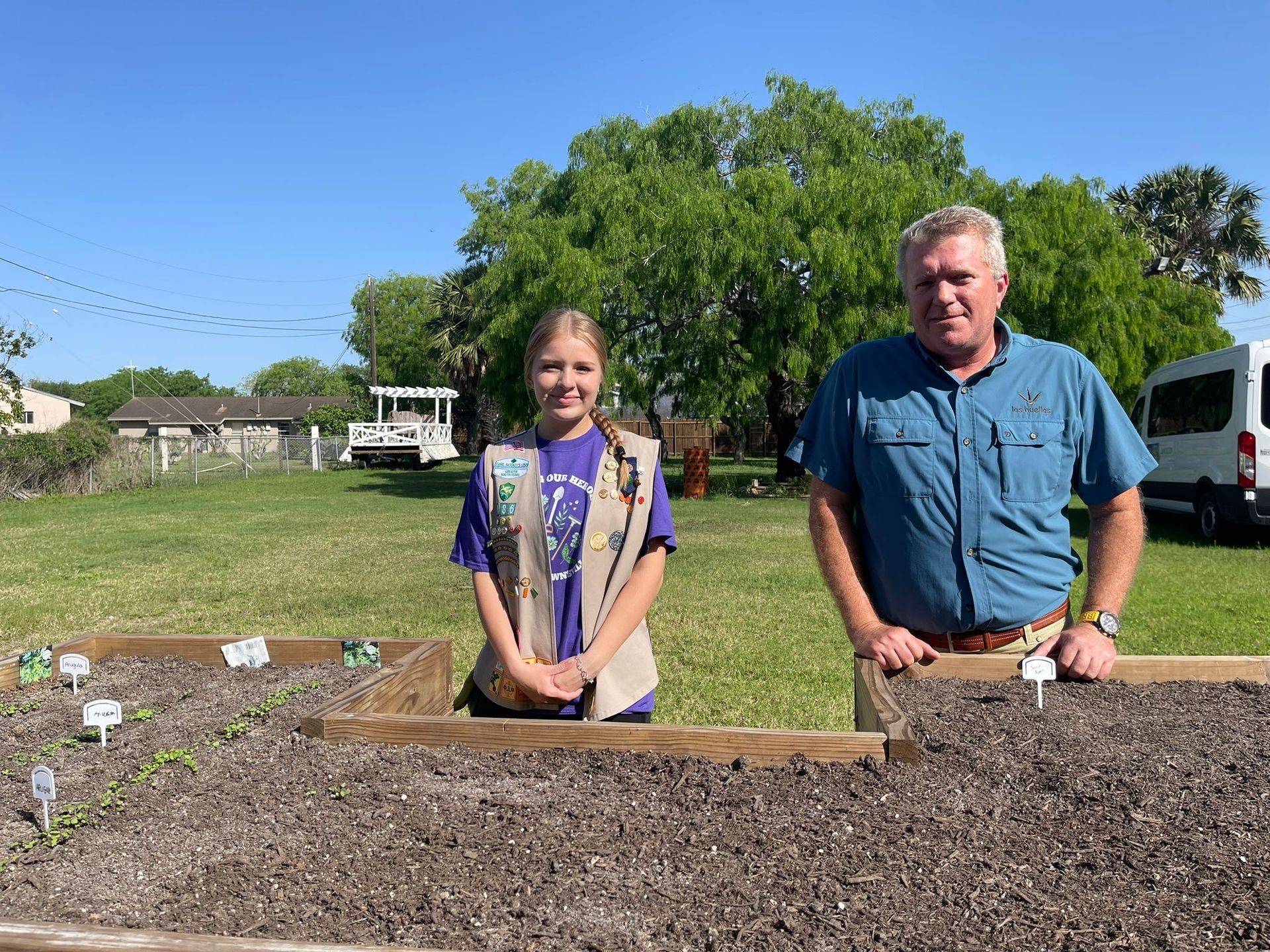 Girl Scout and man stand behind a garden bed with plants, outdoors in a sunny yard.