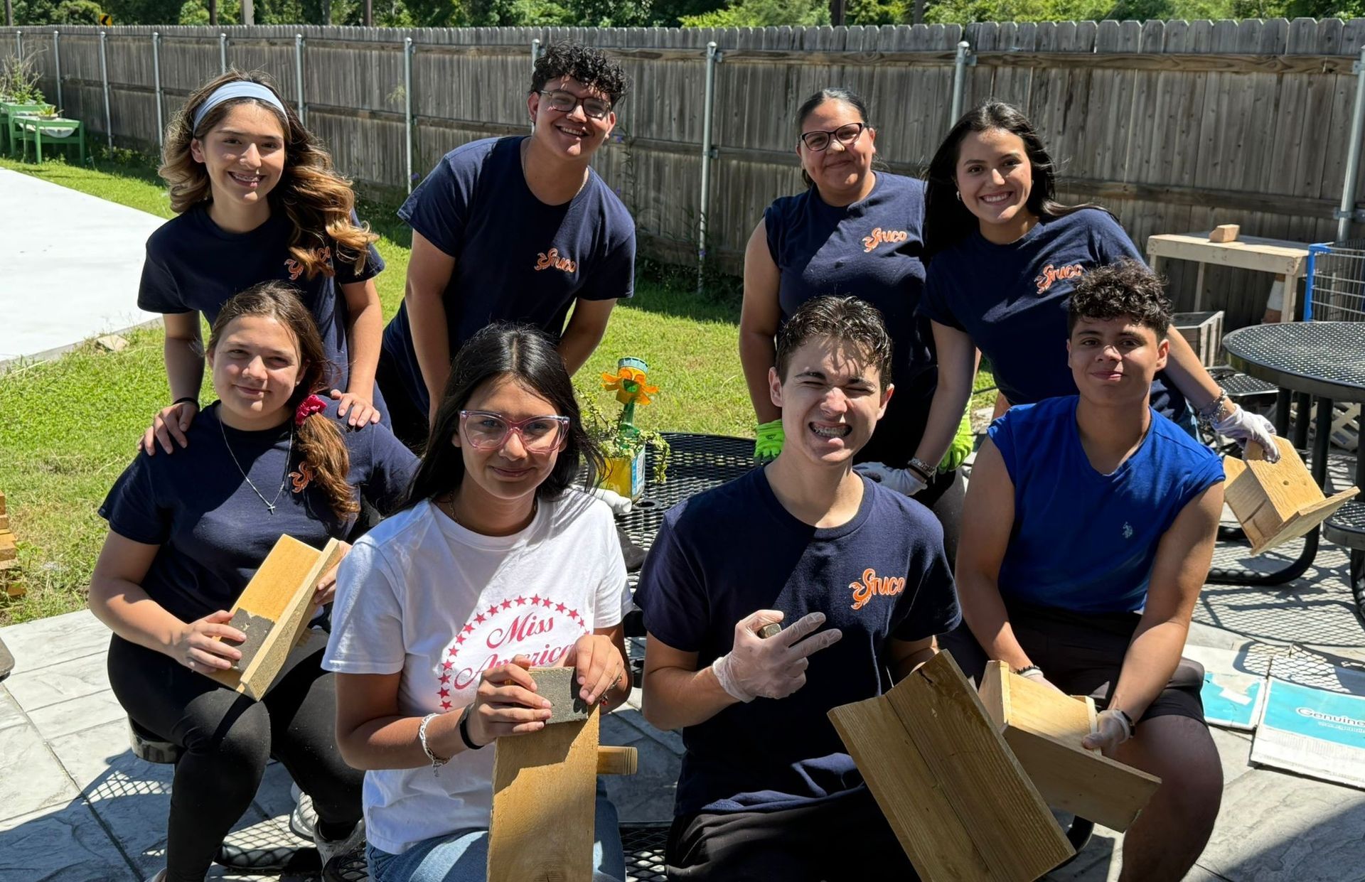 Group of young people in matching shirts, outside, smiling, holding wooden planks, likely volunteering.