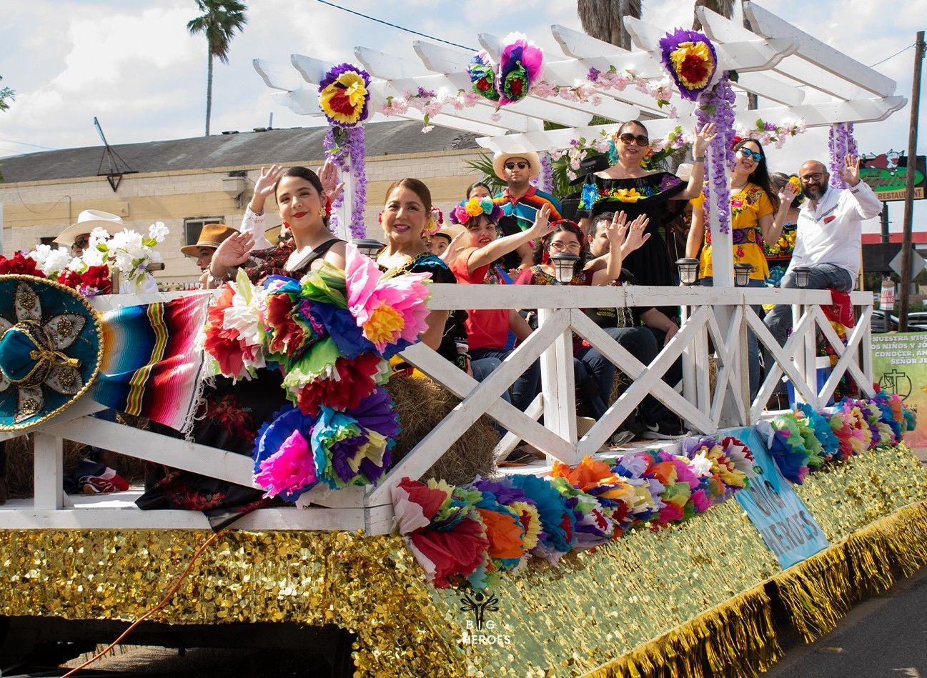 People wave from a colorful parade float decorated with flowers and Mexican flags.