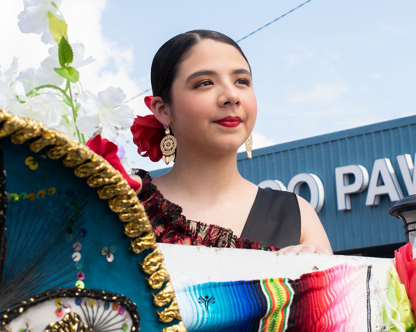 Young woman in traditional dress, red flower in hair, smiling at parade, holding colorful fabric.