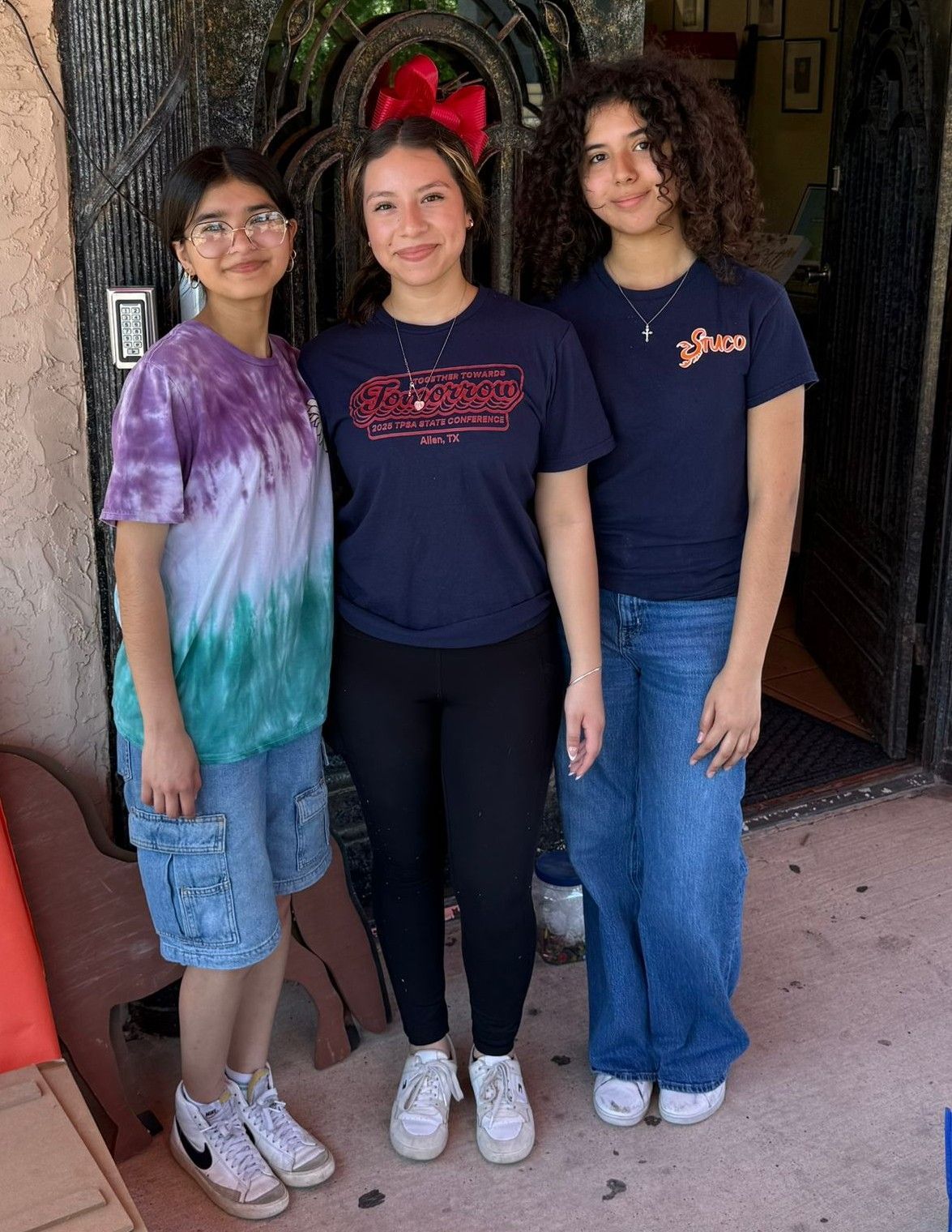 Three young women pose outdoors. One wears tie-dye, blue shorts. Another, black leggings. Third, blue jeans. All smiling.