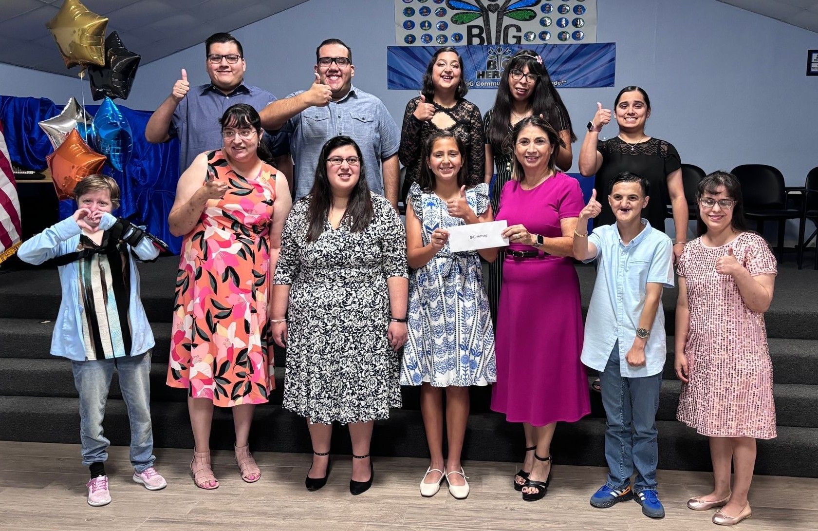 Group of people, some with thumbs up, in a room with a banner. Holding a check, celebratory expressions.