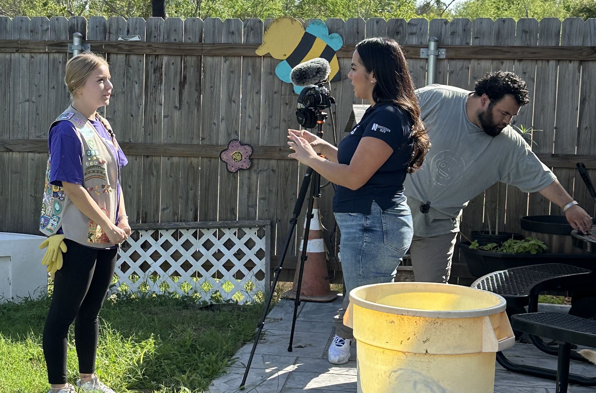 Girl Scout being filmed outdoors by a woman and man near a yellow bucket and wooden fence.