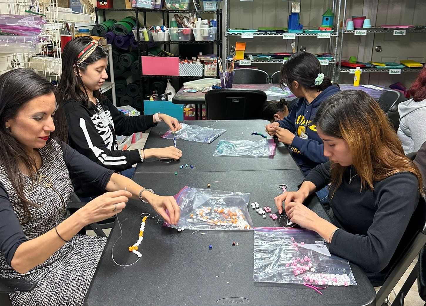 People sitting at a table beading bracelets.  Black clothing, craft supplies, indoor setting.