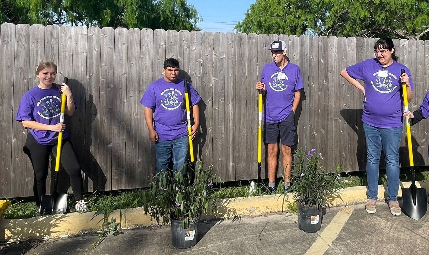 Four people in purple shirts stand next to a fence with shovels, ready to plant trees.