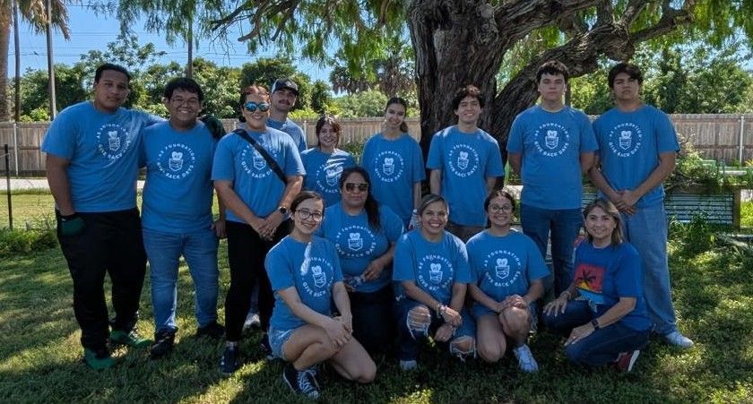 Group of people in blue shirts, smiling, posing outdoors in a sunny garden.