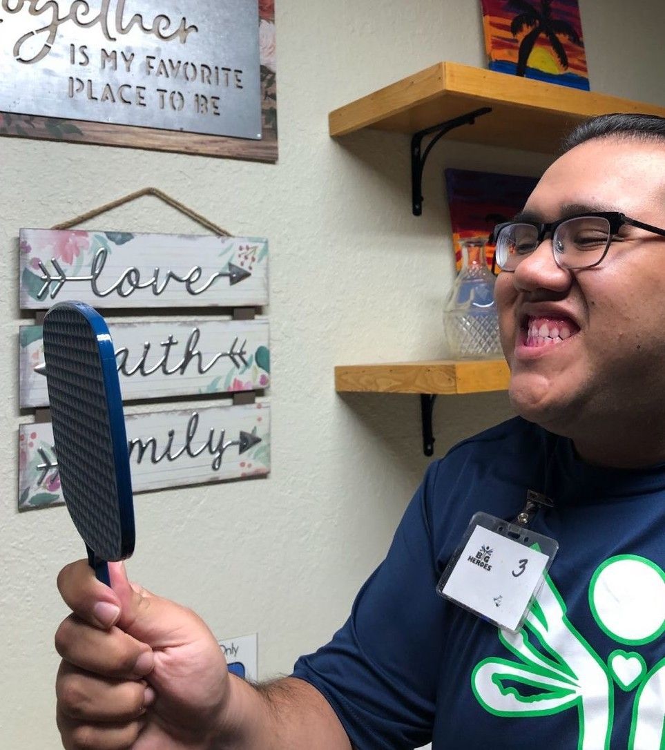 Man holding a blue brush, smiling, near wall with decor. Wearing glasses and a name tag.