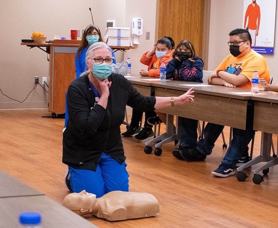 A woman wearing a mask is kneeling down in front of a group of people sitting at tables.