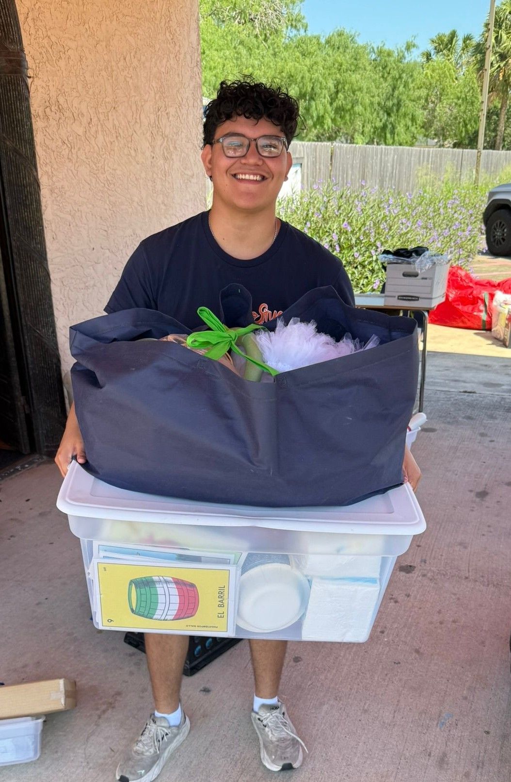 Young man holding a tote bag on top of a clear plastic bin; smiling, outdoors.