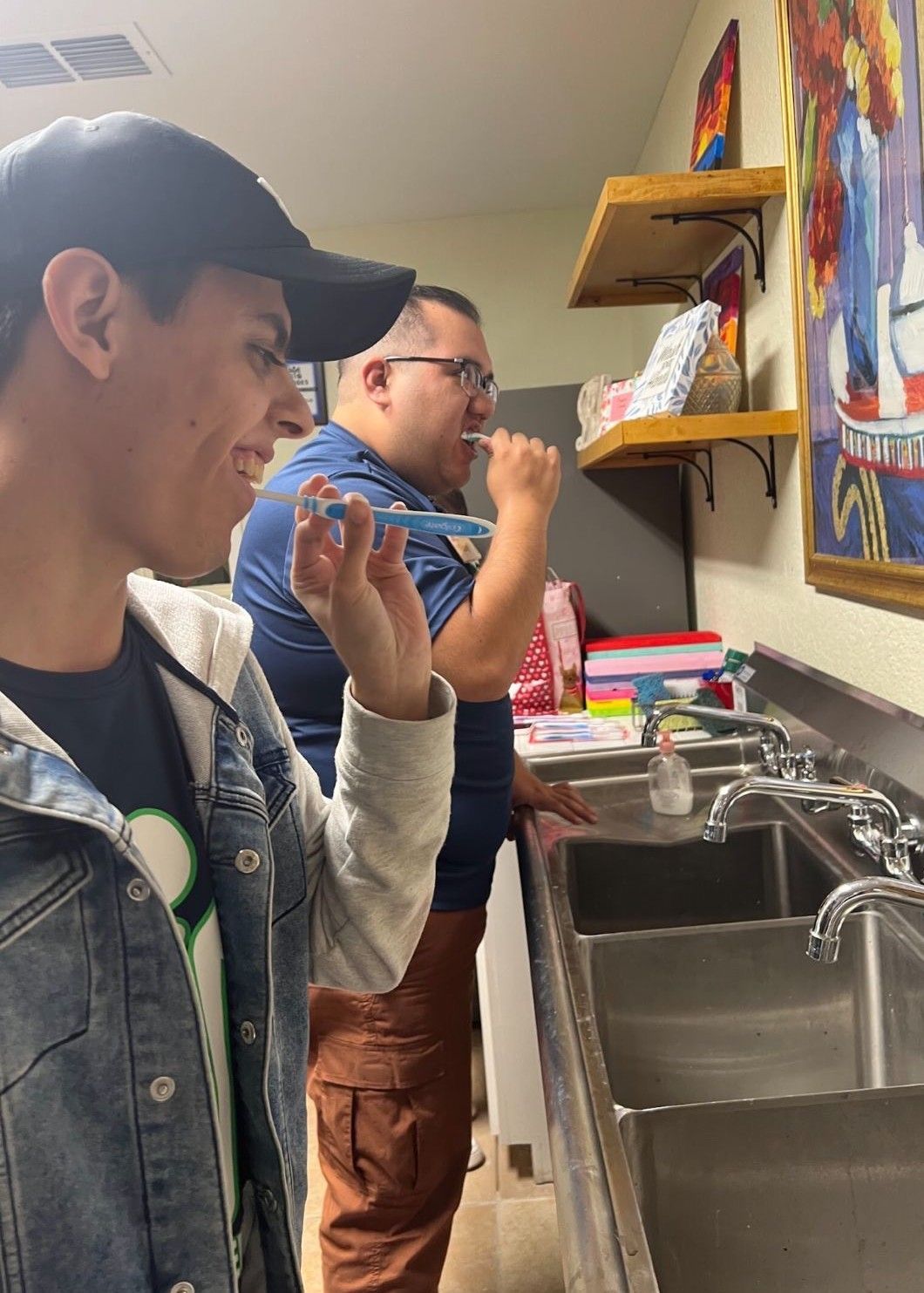 Two people brushing teeth at a sink. One wears a cap, denim jacket; the other, glasses and a blue shirt.