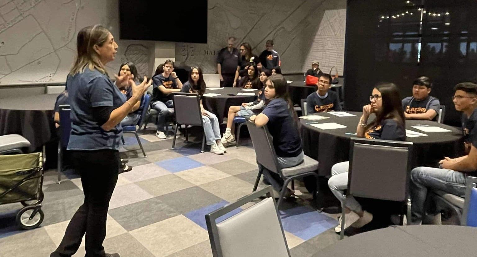 Woman speaking to a group of students in a conference room. They sit at round tables, listening.