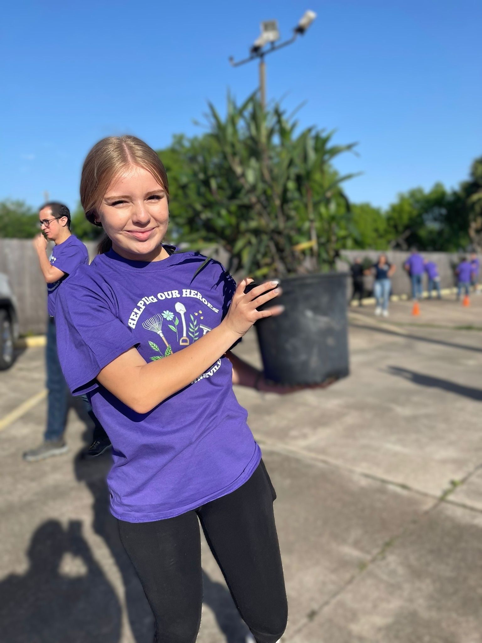 Girl in purple shirt holding a large plant pot, outdoors on a sunny day with others in the background.