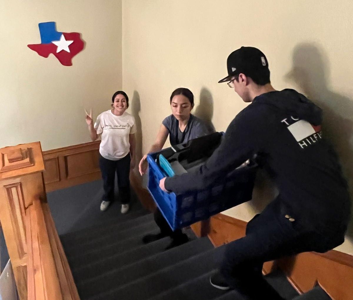 Three people moving boxes on stairs beneath a Texas-shaped decoration.