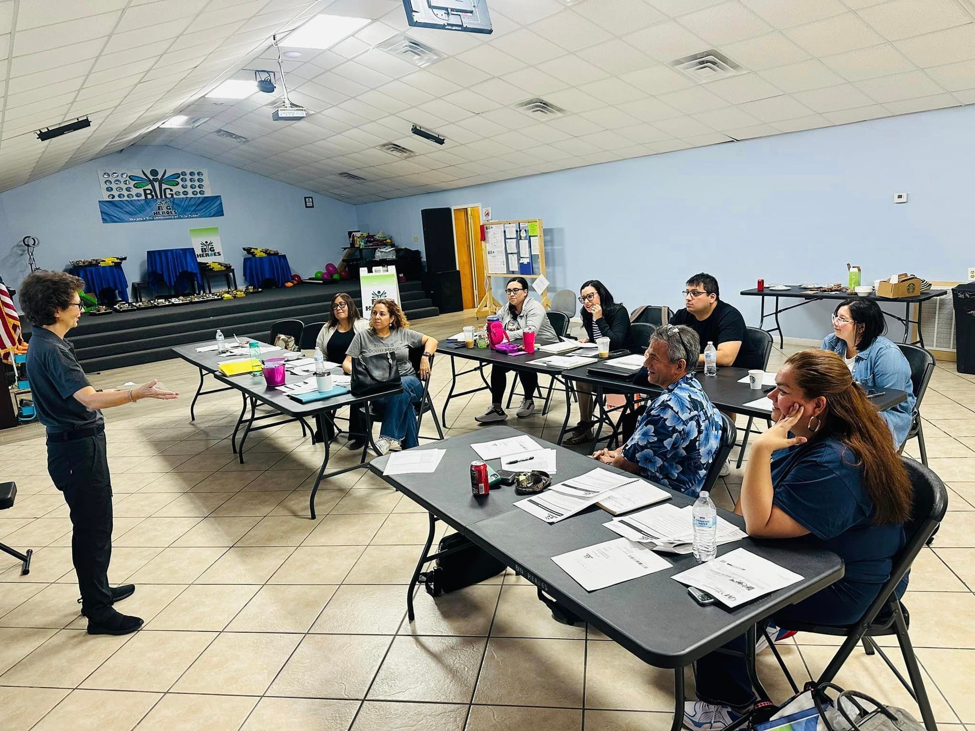 A woman lectures to a group of people seated at tables in a brightly lit room.