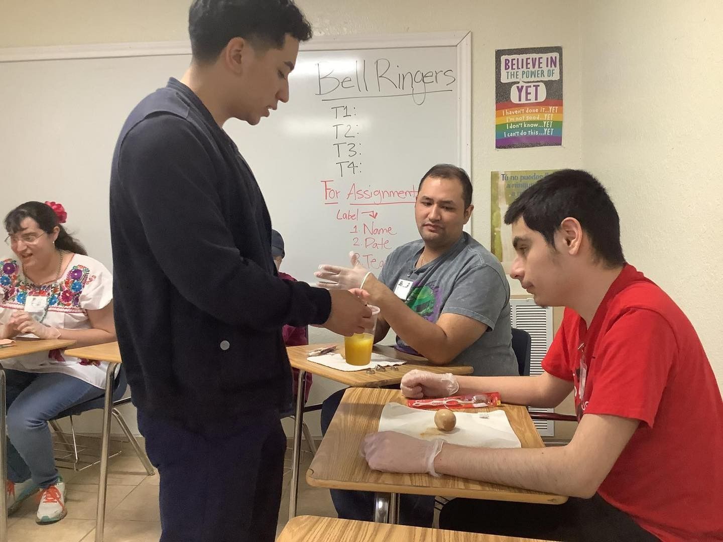 A young man puts gloves on another man in a classroom, where a woman and another man sit.