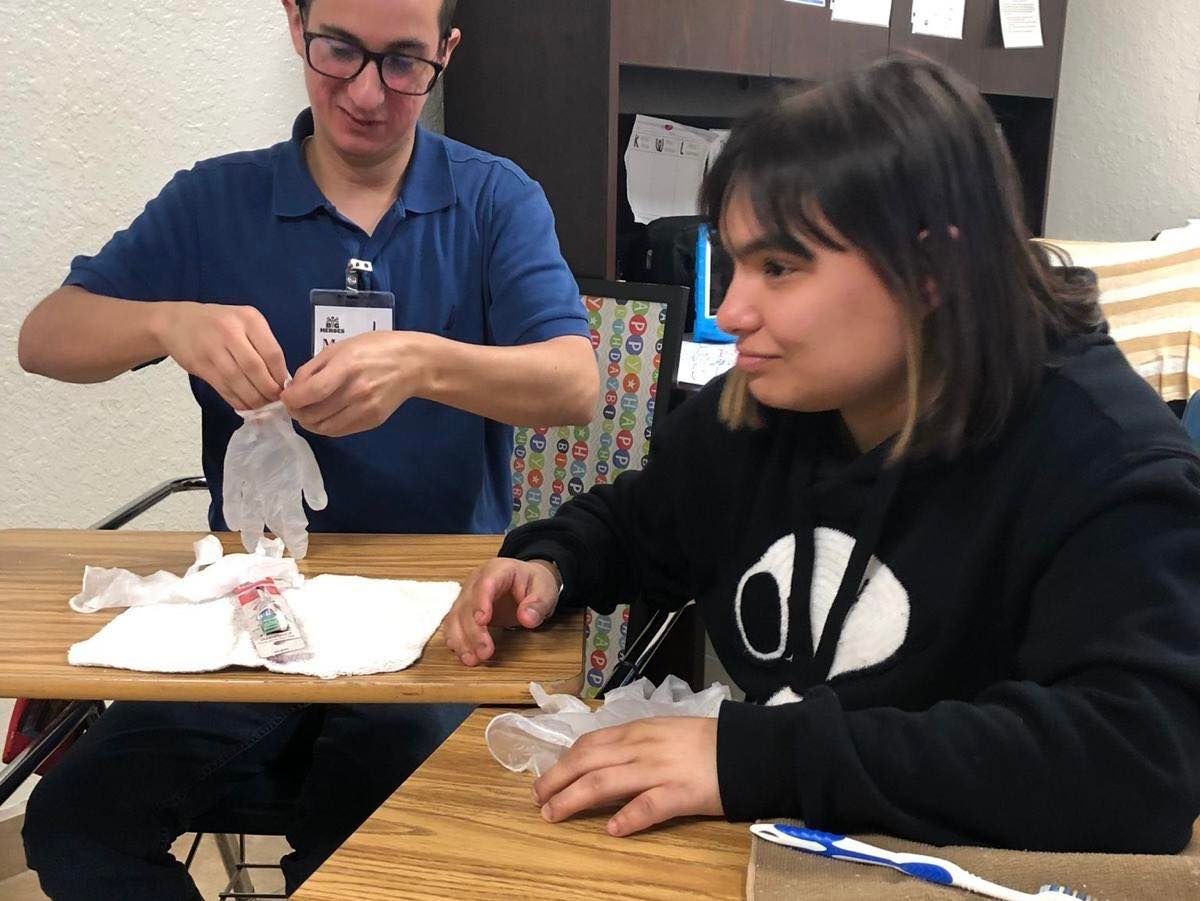 A young man in glasses tying a plastic glove while a young woman watches; both at a table.