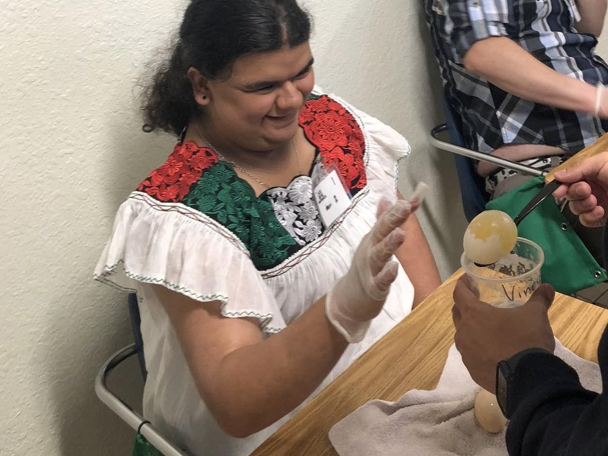 Person in white dress with colorful trim, smiling, reaching towards a cup of liquid being held by another person.